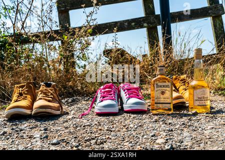 Two bottles, Teacher's Scotch whisky and the Kings Ginger Liqueur, with three pairs of trainers and walking shoes on side of road by a field. Stock Photo
