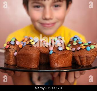 Smiling boy holding a plate of hand-baked Easter cupcakes decorated with colorful candies and sprinkles Stock Photo