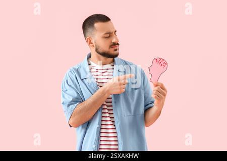 Portrait of thoughtful young man holding paper human head with drawn question marks on pink background Stock Photo