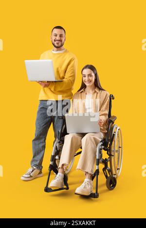 Young woman in wheelchair, with husband and laptops on yellow background Stock Photo