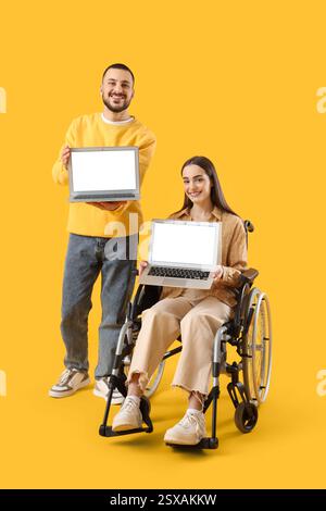 Young woman in wheelchair, with husband and laptops on yellow background Stock Photo