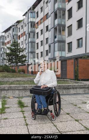 Smiling business lady with disability, a wheelchair user talking on a mobile phone outdoors, in front of the building. Stock Photo