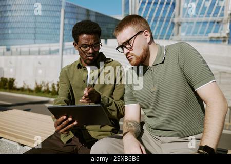 Two young men sitting outdoors, discussing project on a tablet. Background includes modern architectural elements and clear sky, suggesting a productive, open environment Stock Photo