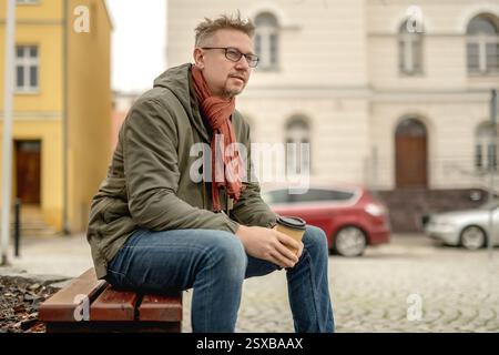 Young Attractive Man Sits On Park Bench Drinking Coffee And Waiting Stock Photo