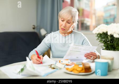 Grey haired thoughtful woman sitting at table and analysing financial documents. She's making notes on paper and counting money. Stock Photo
