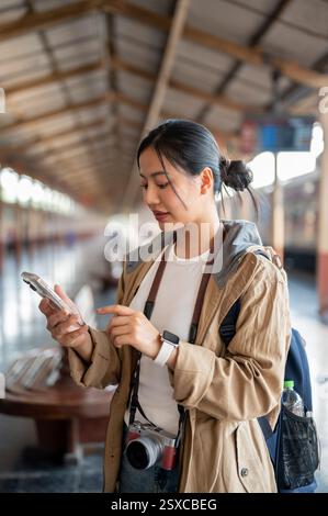 Beautiful asian woman, carrying a camera, is looking through the phone while waiting for the arrival of train at the train station or railway station. Stock Photo