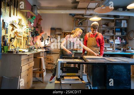 Enthusiastic couple of carpenters prepare stool base for saw machine in workshop. Apprentice teach Stock Photo