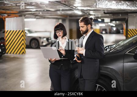 Caucasian male and female business professionals collaborating on laptop in underground parking garage. Man and woman wearing formal attire, emphasizing teamwork, technology Stock Photo