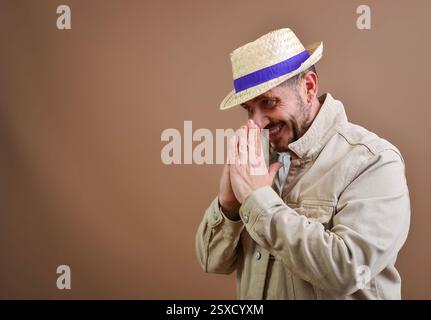 Cunning male tourist wearing casual clothes and a straw hat, rubbing his hands together and smiling, planning something Stock Photo