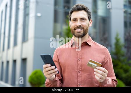 Portrait of happy joyful male shopper with phone in hands and bank credit card. Businessman smiling and looking at camera, walking happily outside office building in city Stock Photo