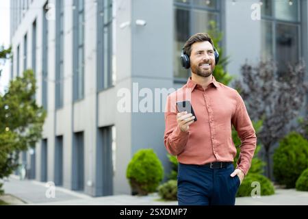 A handsome man walks outside near an office building, enjoying music on his headphones while using his smartphone Stock Photo
