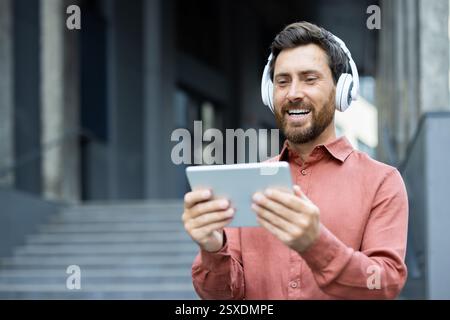 Close-up of a joyful man watching online video while walking outside an office building. Businessman wearing headphones and holding a tablet computer, smiling happily. Stock Photo
