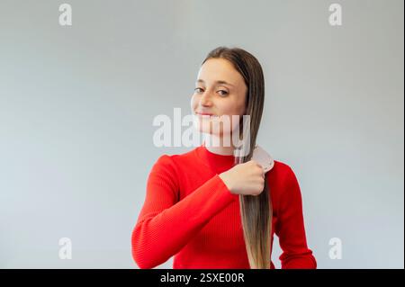 Young woman combing her long hair with comb, wearing red sweater Stock Photo
