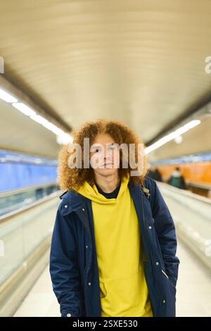 Young man with curly hair wearing a yellow hoodie and blue jacket walking through a brightly lit subway corridor, conveying a sense of urban life Stock Photo