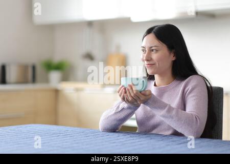 Relaxed asian woman drinking and contemplating in the kitchen at home Stock Photo