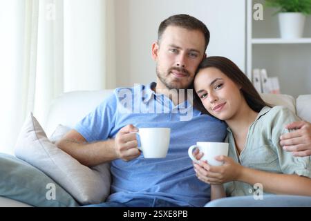 Relaxed couple sitting on a sofa and drinking coffee looking at you at home Stock Photo