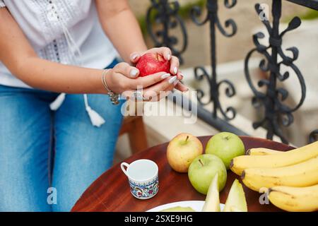 person holds a red apple while sitting on a balcony, surrounded by a selection of fruits such as bananas, green apples, and pears on a wooden table. s Stock Photo