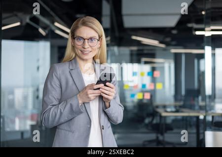 Blonde businesswoman with glasses smiles while holding her smartphone in a modern office setting, ready to tackle the workday Stock Photo