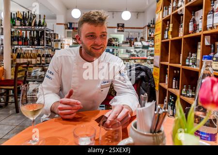 Anton Karnbaum, chef and sommelier, sits at a table during the carp tasting in the wine shop in Forchheim, a glass of white wine in front of him. Apothekenstraße, Forchheim, Bavaria, Germany. Stock Photo