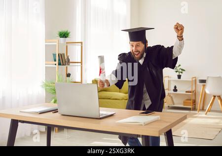 Happy man in student cap and gown celebrating graduation at home after completing online degree Stock Photo