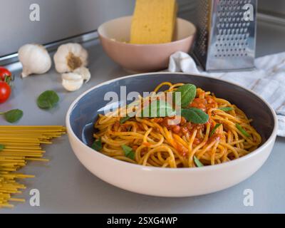 Balanesi pasta gleaming with vibrant tomato sauce, fresh basil scattered, raw spaghetti and garlic surrounding plated dish on textured gray surface Stock Photo