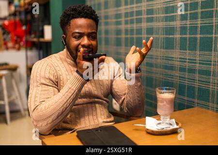 Young man is having pause from work, he is talking on smartphone and drinking hot chocolate in bar. Stock Photo