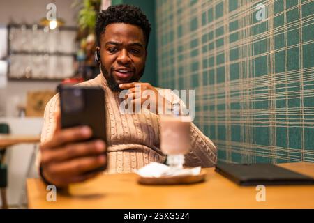 Pensive young man chatting online in bar. He is using smartphone, having video call and drinking hot chocolate. Stock Photo