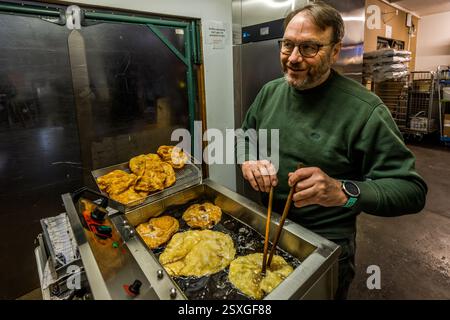 A baker smiles while frying Urrädla with two long wooden sticks in a deep fryer in a bakery on Holzstraße, Forchheim, Bavaria, Germany. Urrädla are a Franconian festive pastry made from egg yolk, sour cream and flour, traditionally baked for confirmations, communions, weddings, christenings and church fairs. Stock Photo