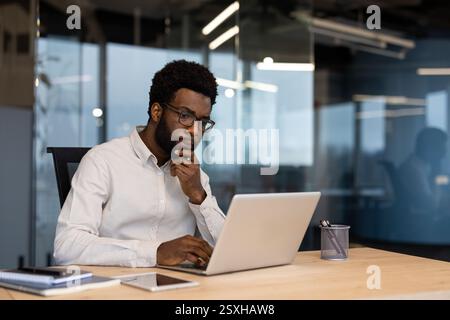 Serious thinking businessman at workplace inside office working with laptop. Man in shirt working concentrated, preparing electronic financial report. Stock Photo
