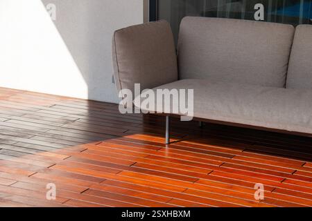 A modern sofa rests on a polished wooden terrace, capturing afternoon sunlight. The soft fabric contrasts with the rich wood tones, creating a cozy ou Stock Photo