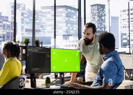 Programmer doing software debugging on green screen PC, asking for help from coworker, checking errors in lines of code. Employee fixing coding mistakes on mockup computer, being assisted by colleague Stock Photo