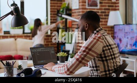 African american software developer works on programming code on a laptop in remote workspace. Productive IT engineer working on coding, software design and internet security innovation. Camera B. Stock Photo