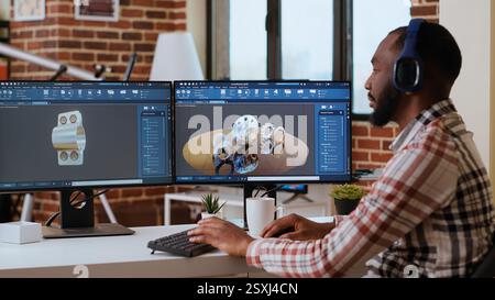 African american industrial designer works on CAD software and listens to music, multitasking at home. Engineer specialist uses digital tools and 3D modeling for industrial projects. Camera B. Stock Photo