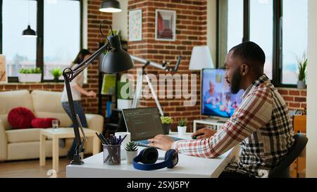 African american software developer works on programming code on a laptop in remote workspace. Productive IT engineer working on coding, software design and internet security innovation. Camera A. Stock Photo