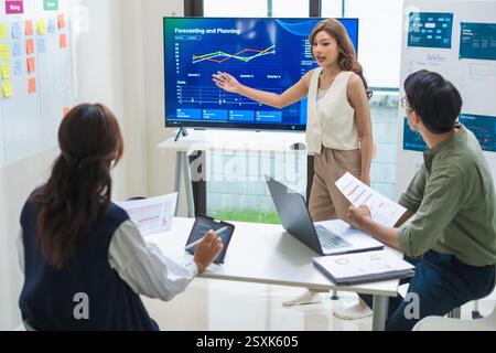 asian leader presenting a financial data and strategies in a modern office with charts and graphs displayed on a large screen Stock Photo