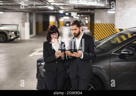 Caucasian man and woman in business attire using smartphones indoors near parked cars. Modern, professional environment with focus on connectivity and communication. Stock Photo