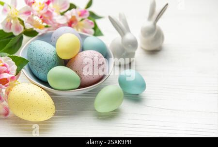 Colorful pastel easter eggs in a bowl with spring flowers and porcelain bunnies celebrating easter holiday on white wooden background Stock Photo