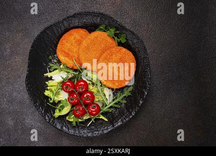Crispy fried fish cutlets, fish burgers, on a black plate, with fresh lettuce leaves, arugula and cherry tomatoes, homemade, no people Stock Photo