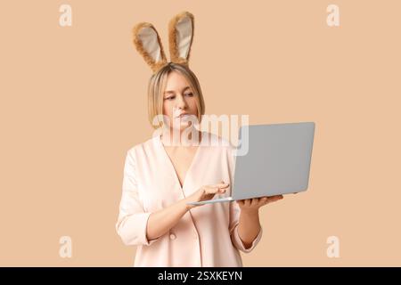 Adult businesswoman in bunny ears with laptop on beige background. Easter celebration Stock Photo