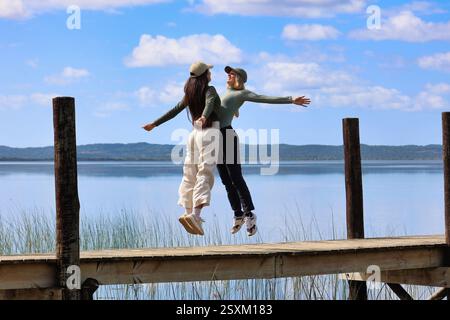 two happy young women jumping on a pier smiling in front of a lake, south africa Stock Photo