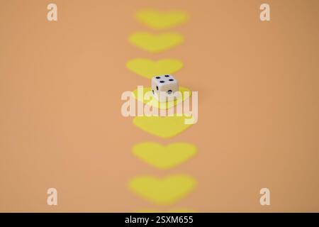 A white dice placed amidst yellow heart shapes creates a symbolic and conceptual arrangement on a warm orange surface Stock Photo