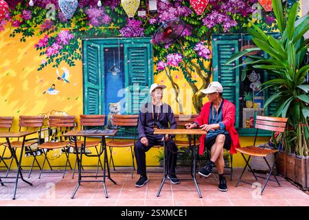 Two men, wearing baseball caps, sitting in an open-air restaurant in Train Street, the wall behind them is decorated with colourful graffiti. Stock Photo