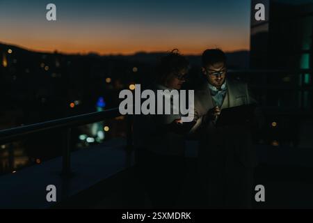 Two business people collaborate outdoors at night, using a tablet for brainstorming ideas. Stock Photo