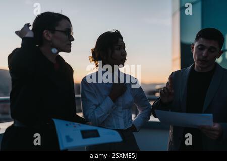 Three business people are engaged in a discussion on a rooftop at sunset, sharing documents and ideas. Stock Photo