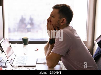 tired young man in a blue shirt is sitting at the desk in the office and working overtime at the laptop Stock Photo