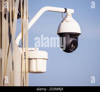 A white dome-shaped security camera with a black lens is attached to a metal arm on the side of a modern building, monitoring the area against a clear Stock Photo