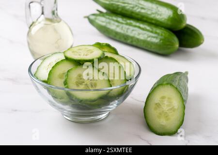 Green cucumber slices in a glass bowl. Long cucumbers, cut in half and bowl of slices over marble countertop. Vegetarian salad, organic vegetables. Stock Photo