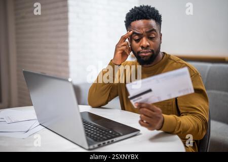 Stressed and depressed young black man using laptop and credit card to pay bills while sitting at his home. He is having financial problems. Stock Photo