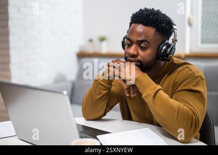 Tired and bored young African-American man with wireless headphones struggling to concentrate while attending online meeting on laptop at his home. Stock Photo