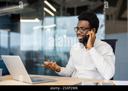 Successful man talking on the phone at workplace while sitting at desk with laptop. Businessman is concentrated in negotiations with colleagues and partners. Stock Photo